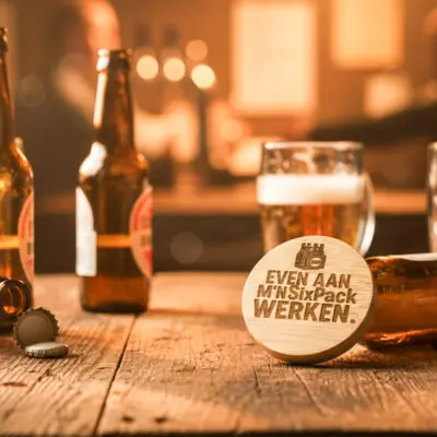 Ronde houten bieropener met gegraveerde tekst “Even aan mijn sixpack werken”, gemaakt van bamboe met metalen onderzijde. Ambachtelijk gegraveerd door De Meesterbrander.