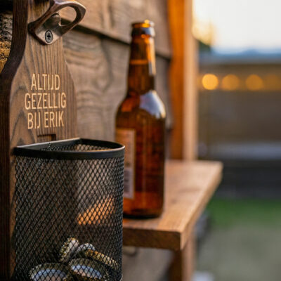 Afneembaar metalen dopjesmandje gevuld met bierdopjes bij houten wandbieropener