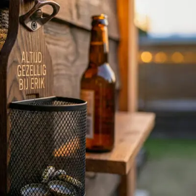 Houten Wandbieropener met Dopjesmandje Afneembaar metalen dopjesmandje gevuld met bierdopjes bij houten wandbieropener