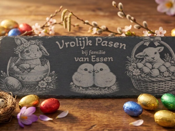 Rechthoekige leisteen met gravure van konijn, kuikens en lammetje omringd door kleurrijke paaseitjes op een houten tafel.